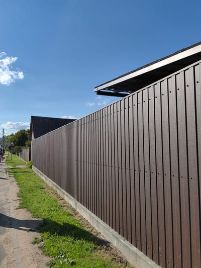 This image captures a scene on a sunny day in Fanipal, Belarus. The foreground is dominated by a tall, dark brown corrugated metal fence that runs along the right side of the frame, extending into the distance. A concrete base supports the fence, with a strip of green grass growing along its edge. To the left of the fence, a paved sidewalk is visible, with a dusty dirt path bordering it on the far left. A shadow of a person is cast on the sidewalk, suggesting it is late morning or early afternoon.
Further down the sidewalk, a few people are walking. One person is closer to the viewer and appears to be walking away from the camera, with another person partially visible behind them. A triangular road sign with a red border is planted in the grass near the sidewalk, with a second, less distinct sign visible behind it. Beyond the fence and the sidewalk, a sloped, dark-roofed structure, possibly a house or shed, is partially visible. Lush green trees form a backdrop to this structure and the fence.
The sky is a clear, bright blue with a few scattered white clouds on the left side of the frame. The top right corner of the image shows a portion of a dark, angled structure, possibly a roof or awning, with metal supports. The overall impression is one of a quiet, residential or semi-rural area on a pleasant, sunny day.
