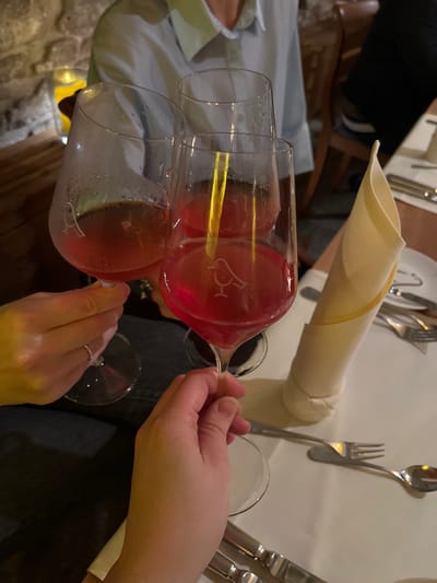 A table is set with silverware, a napkin, and two hands holding wine glasses. One glass is mostly full of a rose-colored liquid and is being poured into a second, nearly empty glass. The wine glasses have a small emblem on them. Two people sit at the table, visible from the chest up.