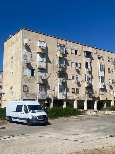 The image shows a white Mercedes-Benz Sprinter van parked on a paved lot in front of a weathered, light-colored, multi-story apartment building under a clear blue sky. The building exhibits signs of age and disrepair, with peeling paint and visible air conditioning units mounted on its exterior. The van is positioned on the left side of the frame, facing slightly towards the right. The apartment building, typical of residential structures in Kiryat Yam, Israel, features numerous windows, some with closed shutters, and balconies. Green bushes and vegetation are visible around the base of the building and in front of some of the ground-level openings. The paved area in front of the building appears to be a parking lot or communal space.
