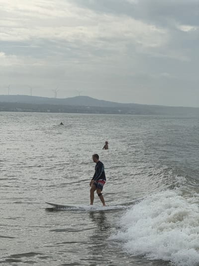 A medium shot captures a surfer in motion on a white surfboard in the ocean near Phan Thiết, Vietnam. The surfer, a male with fair skin, is wearing a dark, long-sleeved rash guard with blue and white striped accents on the sleeves and patterned board shorts. He is positioned in the center of the frame, riding a small wave. His body is turned slightly to the left, and his right leg is forward. To the right of the surfer, a frothy wave is breaking. In the distance, two other individuals are in the water, one appearing to be sitting on a surfboard and the other floating. The ocean is calm with gentle ripples, and the sky is overcast with heavy clouds. In the background, a hazy, mountainous coastline is visible, with several wind turbines dotting the ridge. Buildings are scattered along the coast. The overall lighting suggests it is daytime, likely late afternoon or early morning due to the soft, diffused light.