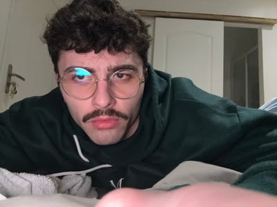 A man with curly dark hair and a fake mustache peers into the camera. He is wearing round glasses and a dark green hoodie. The photo is taken from an upside-down perspective. A white door and window are visible in the background, along with a white towel. His hand is partially visible at the top of the frame.