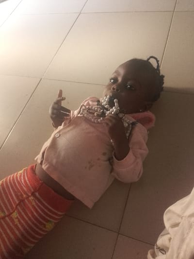 An image taken in Oturkpo, Nigeria, depicts a young child lying supine on a light-colored tiled floor. The child has dark skin and dark hair, styled with a visible loop on top of the head. They are dressed in a light pink long-sleeved top, soiled with several dark stains on the front, and orange and white striped pants featuring yellow patterns. The child is actively holding a cluster of white, spherical beads, likely a necklace or rosary, with both hands, and has several of these beads in their mouth. The child's gaze is directed slightly upwards and to the right. The indoor environment is lit dimly, and a white fabric object is partially visible in the bottom right corner of the frame.