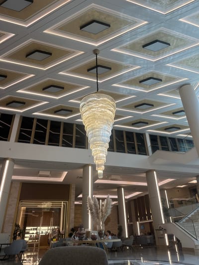 The image shows the interior of a modern, multi-level building, likely a hotel or a large commercial space, with a focus on sophisticated design elements. A large, multi-tiered glass chandelier is the prominent object in the foreground, hanging from the ceiling. The walls feature a decorative pattern of interlocking diamonds, with inset square panels and integrated lighting that creates a warm glow.
The background reveals the building's structure, including multiple floors with balconies or walkways overlooking the central atrium. Large windows and glass railings are visible, reflecting the interior lighting. In the distance, there are glimpses of seating areas, possibly a lounge or cafe, with people seated at tables and chairs. Decorative plants and dried floral arrangements are also part of the interior decor.
The lighting suggests it could be daytime or evening, with the artificial lights playing a significant role in illuminating the space. The overall impression is one of elegance and contemporary style. No specific activity or event is clearly depicted, but the presence of people and seating areas implies a public or commercial function. No text is visible in the image. The provided location context of Kazanlak, Bulgaria, does not offer specific visual cues within the image itself.