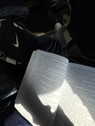 A person is sitting, presumably on a train or bus, reading a book. The person is wearing dark pants with white vertical stripes and white sneakers. The book is open and lying on their lap. The text in the book is in Bulgarian. The book appears to be a medical text, likely concerning childbirth or gynecology. There is a black Nike bag near the left side of the image. The location appears to be indoors in transit, potentially in Byala Slatina, Bulgaria, but this is speculative based on the Bulgarian language in the book.