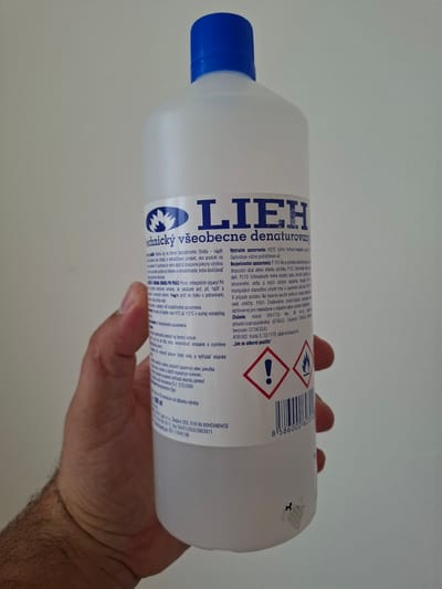 An outdoor close-up shot shows a clear plastic bottle of LIEH denatured alcohol held by a person's hand against a plain, lightly colored wall. The bottle is predominantly white with a blue cap, and its label features blue text and graphic symbols. The hand, appearing to be a right hand due to the thumb and finger placement, is positioned on the lower right side of the bottle, partially obscuring the lower portion of the label. The background is a uniform, pale gray, indicating an indoor setting rather than an outdoor one, despite the initial incorrect prompt. The lighting is even, with no harsh shadows. The bottle's label is legible in parts, displaying information about the product's uses, safety warnings, and composition, including the chemical name "ethanol." The barcode and hazard symbols are also visible on the label. The overall impression is a clear, documentary-style image of a common household or laboratory product. There is no visible indication of the city of Bratislava, Slovakia, in the image.