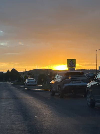 A wide shot captures a rural road in Большой Кукмор, Russia, at sunset. The sky is a vibrant blend of orange and yellow, with wispy clouds streaking across the horizon. A line of trees and hills forms the backdrop. In the foreground, a paved road is visible with two lanes marked by white lines. Several cars are driving on the road; a white sedan is in the left lane, and a darker SUV is in the right lane. The SUV has a license plate that reads "750DCX777". A yellow triangular road sign with a pedestrian symbol is mounted on a pole to the right of the SUV. The scene is illuminated by the warm glow of the setting sun, casting long shadows across the road. The overall impression is one of peaceful, rural life at the end of the day.