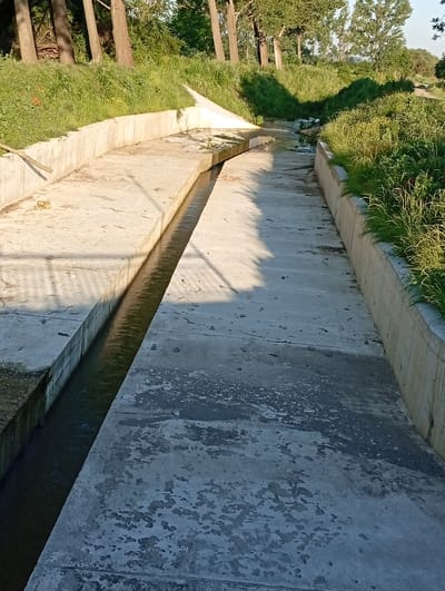 The image depicts an outdoor scene in Popovo, Bulgaria, featuring a concrete channel with a narrow stream of water flowing through it. The channel is lined with concrete walls, and the surrounding area is covered in lush green grass and trees, suggesting a natural or semi-natural environment. The lighting indicates it is daytime, with shadows cast on the concrete surface. There are no people visible in the image, and the overall atmosphere is one of quietude and natural beauty.