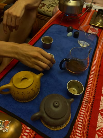 The image depicts an indoor scene focused on a tea preparation or serving ritual. A person's light-to-medium skin-toned hands are prominent, with the right hand holding a small white teacup in the process of being placed or picked up. The main activity occurs on a blue microfiber mat, which overlays a red, white, yellow, and black geometrically patterned fabric runner.

In the upper-right background, a silver metal kettle rests on a small, three-legged stand, with a visible blue flame heating its underside. Behind it, a small dark reddish-brown cup is visible, and to the right, a golden tray holds several clear empty glasses and one dark cup.

Arranged on the blue mat are various tea implements:
- A mustard-yellow clay teapot, adorned with embossed or carved Chinese characters, rests on a woven coaster in the lower-left quadrant.
- A dark brown clay teapot, simpler in design, also on a woven coaster, is positioned to the right of the yellow teapot.
- A clear glass serving pitcher with a black handle contains an amber-colored liquid, presumed to be tea, positioned in the mid-right.
- A small, empty dark brown cup with a lighter rim is situated below the glass pitcher.
- Three decorative, smooth stones are centrally placed: two dark, spherical stones and one light blue, irregularly shaped stone.
- An empty, clear glass with a distinctive angular, V-shaped rim is visible in the upper-right corner of the blue mat.

In the upper-left background, parts of a person's upper arm and leg are visible, along with a portion of wicker furniture and a cream-colored cushion with a floral pattern. A golden container with a sticker depicting an octopus-like creature holding a weapon is visible in the lower-left foreground. The image provides no visual information to determine its location as Zima Yuzhnaya, Russia.