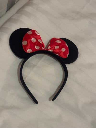 A Minnie Mouse ear headband lies on a white, textured surface. The headband is black and slightly curved. The ears are black circles, adorned with a red and white polka dot bow in between them. The bow is made of shiny fabric. The surface appears to be a bed or a similar soft furnishing.