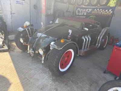 The image shows a custom-built car with skull decorations, large wheels, and multiple exhaust pipes, parked in what appears to be a garage or workshop setting. A Coca-Cola vending machine, tools, and signage (Sharko Zic, Crazy Cars Show on Wheels) are visible in the background. While Općina Funtana, Croatia is not explicitly identifiable, the car's style and the environment suggest a custom vehicle shop or exhibit location, possibly related to automotive events or displays.