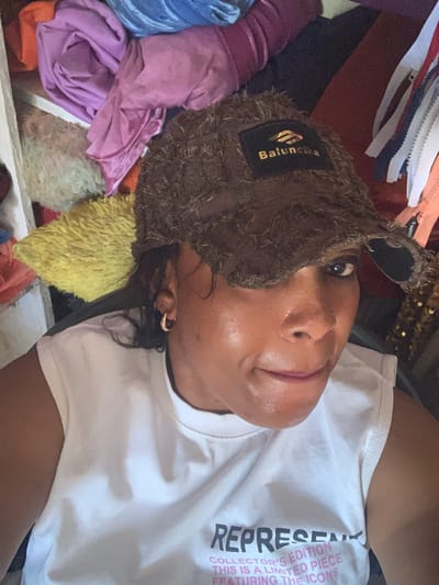 The image depicts a woman wearing a distressed brown baseball cap and a white sleeveless t-shirt with text that reads "REPRESENT COLLECTOR'S EDITION THIS IS A LIMITED PIECE FEATURING THE ICONS." The background is cluttered with various fabrics, including purple, blue, and yellow materials, as well as zippers and some sequined fabric. The lighting suggests an indoor environment. There is no information in the image that would indicate the location is Yelwa, Nigeria.
