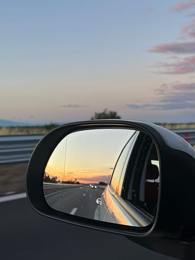 The image shows a side-view mirror of a car reflecting a sunset over a highway. The sky is a gradient of blue, pink, and orange. In the distance, there are faint outlines of mountains and trees. The highway is visible, with lane lines and other cars in the reflection. The car itself is white, and its interior is partially visible through the window. The image is taken from a high angle, looking down on the mirror. There are some water droplets on the mirror's surface. The location is identified as Malo Konare, Bulgaria.