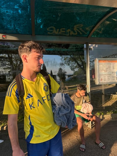 Two men are at a bus stop in Rijeka, Croatia. One man, in a yellow and blue soccer jersey, stands facing left, with a dark backpack, while the other man is sitting on the bus stop bench, wearing a brown and green shirt and green shorts, with a pink towel on his head. He is looking at his cell phone. He is wearing a pair of black and white Adidas slides. A blue sweater is on the bench next to him. The bus stop has a clear glass back with information on it. The green marble glass on the side of the bus stop has the word 'SUENTLY' in yellow writing. A sign underneath the glass contains the words, 'GRAD LIEKA.' The scene appears to be outside, with trees and a building reflecting in the bus stop glass.