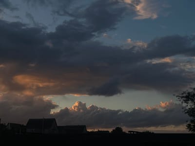 The image depicts a dramatic sky at dusk over a rural landscape in Volodarka, Ukraine. Dark, heavy clouds dominate the upper portion of the frame, with streaks of golden and pink light piercing through them, suggesting the last rays of the setting sun. The lower section of the image is silhouetted against the illuminated sky, revealing the dark outlines of houses with steeply pitched roofs. A few trees are also visible as dark shapes against the horizon. The overall scene conveys a sense of impending twilight and the raw beauty of nature.