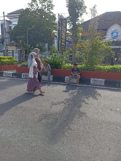 This outdoor image captures a street scene in Mataram, Indonesia, under clear skies with strong sunlight, indicated by the long shadows cast across the asphalt road, suggesting it is either morning or late afternoon.
In the foreground, a paved road is bordered by a curb painted with distinct black and white stripes. Two women are walking across the road from left to right. The woman in front wears a light grey hijab, a white top, and wide-legged purple pants, with sandals on her feet. Behind her, another woman is dressed in a brown hijab and a darker top, carrying a bag.
Along the curb, two men are seated, facing towards the right side of the frame. The man on the left wears a striped short-sleeved shirt and jeans. The man on the right is dressed in a dark t-shirt, shorts, and a cap, and appears to be holding a mobile phone.
Behind the seated men and the curb is a neatly maintained garden area with various green plants and small trees, contained within a raised planter with red and black borders. In the midground and background, several tall trees are visible, along with multi-story buildings. A prominent vertical billboard or banner stands tall, featuring a black background with yellow and white text, which appears to include "SELAMAT DATANG" (Welcome) in Indonesian. One building in the background on the right displays a circular blue and white emblem on its facade. Another individual in a dark shirt can be seen walking further in the distance on the right. The scene depicts everyday activity in a sunny urban setting.