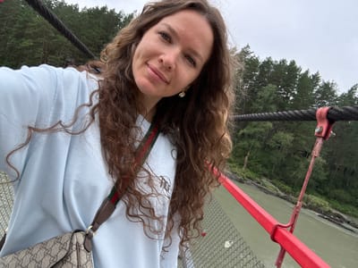 A woman with long, curly brown hair takes a selfie on a suspension bridge. She is wearing a light blue sweatshirt and a designer shoulder bag. The bridge has thick cables and red metal railings. In the background, a river flows through a forest. The image appears to be taken in Barangol, Russia, a location known for its scenic suspension bridges and natural beauty. The sky is overcast, suggesting a cloudy day. The woman is smiling and looking directly at the camera. The overall scene depicts a tourist enjoying the landscape.