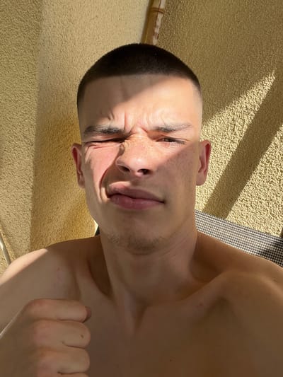 A close-up, low-angle shot depicts a shirtless man with a short, dark haircut, lying on a textured surface. The man is squinting, his face contorted in a grimace. Light from the sun falls across his face, casting shadows. His hand, balled into a fist, is visible near his shoulder. The background consists of a beige, textured surface and a glimpse of a patterned object. The location cannot be determined by visual inspection of the provided image. Sopot is a city in Poland, not Bulgaria.