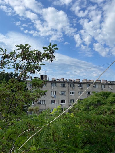 The image captures a view of a residential apartment building in Yevpatoriya, Ukraine, partially obscured by lush green foliage from trees in the foreground. The building is a multi-story structure with a grey facade, featuring numerous windows and air conditioning units mounted on the exterior. The rooftop is adorned with multiple chimneys. The sky above is a bright blue, dotted with fluffy white clouds. In the foreground, branches of a tree with green leaves and brown seed pods extend into the frame, with a thick white cable running diagonally across the lower right portion of the image. The overall scene suggests an urban setting with significant greenery.