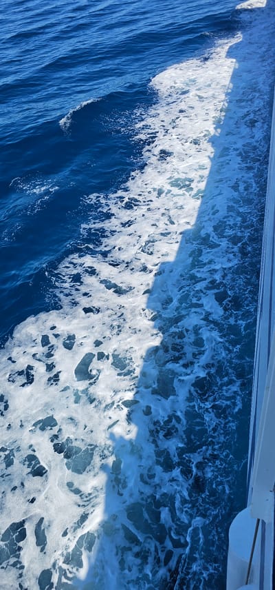 This is a high-angle, vertical shot of a boat's wake on the sea, seen from the perspective of someone on board. The water is a deep blue and frothy white, with the white foam churned up by the boat's passage creating a distinct line separating the calmer water from the turbulent wake. A white railing or side of the boat is visible on the right edge of the frame, casting a long shadow across the water. The scene appears to be set on the sea near the city of Supetar, Croatia.