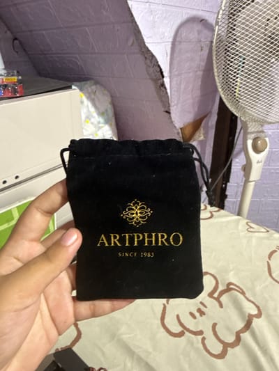 A person's hand is holding a black velvet pouch with gold lettering that reads "ARTPHRO SINCE 1983". The pouch is slightly open, revealing some of its contents. In the background, there is a white fan with a mesh grille, a wall with textured purple wallpaper, and a cream-colored surface with a patterned fabric draped over it. The overall scene suggests an indoor setting, possibly a room in a home. The lighting appears to be artificial, with no strong indicators of the time of day or weather. The focus is on the pouch and the hand holding it.
