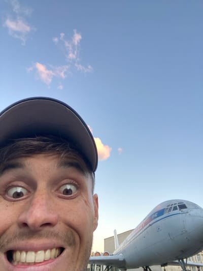 The image is a selfie taken by a man with wide eyes and a large smile. He is wearing a dark baseball cap. Behind him, to the right, is a white airplane with blue and red stripes. The text "ИА-62" is visible on the side of the airplane. The sky is blue with a few pink clouds. The airplane appears to be on display, possibly at a museum or outdoor exhibition. The background suggests an urban environment with buildings visible. The overall impression is of a playful or enthusiastic pose in front of an aircraft. While the image is taken outdoors, the specific location within Ulyanovsk, Russia cannot be determined from the visual information alone.