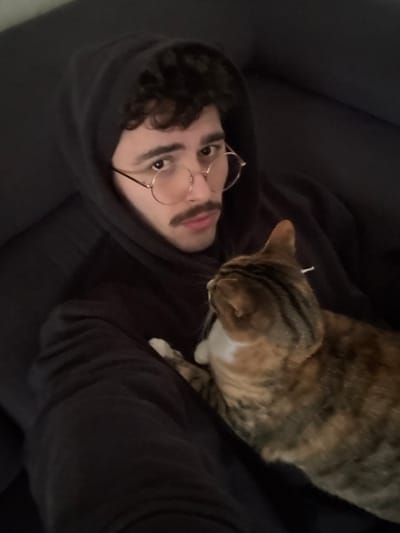 An indoor scene depicts a young adult male with dark, curly hair, a mustache, and round, gold-rimmed glasses. He is wearing a dark hooded sweatshirt with the hood up, and is seated against a dark upholstered surface, possibly a couch or armchair. He looks directly into the camera with a neutral expression. A domestic short-haired cat with a calico or tortoiseshell coat (brown, orange, and white patches) is resting on his lap or chest, facing away from the camera and towards the viewer's right. The cat's white chest and front paws are visible. The specific location in Rennes, France, cannot be determined from the visual information provided in the image.
