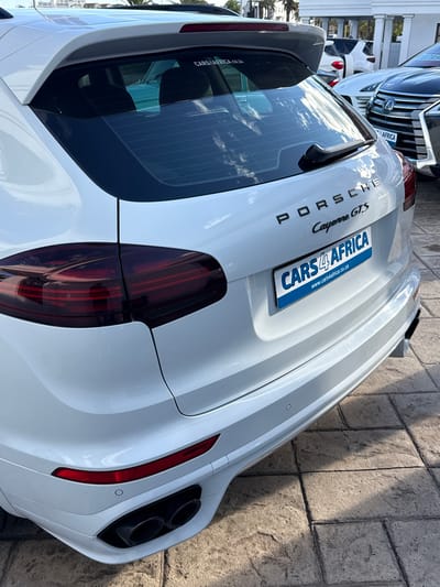 The image displays the rear of a white Porsche Cayenne GTS SUV, parked outdoors on a light-colored tiled surface during bright daylight. The sun casts distinct shadows on the ground, and reflections of clouds and surrounding buildings are visible on the car's glossy surface and rear window.
The prominent features of the car include the "PORSCHE" lettering and "Cayenne GTS" badge on the tailgate. A blue and white plate is affixed to the tailgate, bearing the text "CARS 4 AFRICA" and "www.cars4africa.co.za". Another sticker on the lower left of the rear window reiterates "CARS4AFRICA.CO.ZA". The vehicle is equipped with a rear wiper, distinctively shaped red LED taillights, and dual large black exhaust tips on the right side of the rear bumper. Small circular parking sensors are also visible on the bumper.
In the background, several other parked cars are partially visible, including an SUV with a large front grille and a license plate that also contains the word "AFRICA". The scene suggests a car dealership or a parking lot, possibly in Blouberg, South Africa, given the contextual information and the ".co.za" domain on the visible text. No people are present in the image.