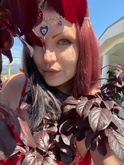 A portrait-style shot captures a young woman with reddish-brown hair and fair skin, positioned outdoors in Kunje, Montenegro. She is looking directly at the camera with a calm, neutral expression, a slight smile playing on her lips, and her green eyes are prominent.
The woman's head is tilted, and her face is partly framed and partially obscured by numerous dark, deep red-purple leaves belonging to a nearby plant or branch. Small, dark red berries are also visible among these leaves in the foreground. Sunlight highlights the textures of the leaves and parts of her hair.
She is adorned with intricate accessories. A delicate gold-colored chain with a small heart charm is visible near her temple. Below her eye, a larger, ornate pendant hangs, featuring an 'evil eye' motif within a red and silver heart design, embellished with rose-like details. Her makeup includes dark eyeliner and mascara, emphasizing her eyes.
A red garment, possibly a top or dress with a subtle lace pattern, is visible on her shoulder and chest. The background features a clear blue sky and a white architectural structure, suggesting a bright, sunny daytime setting. The overall composition creates an ethereal and somewhat mysterious mood, with the dark leaves contrasting with her skin tone and the bright sky. No visible text is present in the image.