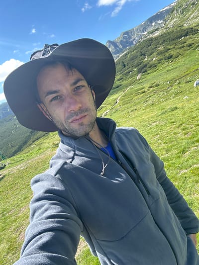 A man wearing a wide-brimmed hat and a grey fleece jacket is taking a selfie on a grassy mountain slope under a clear blue sky with scattered clouds. The background features a lush green mountain covered with sparse trees, with a jagged mountain peak visible in the distance. The man's gaze is directed towards the camera. The scene is set in Panichishte, Bulgaria.