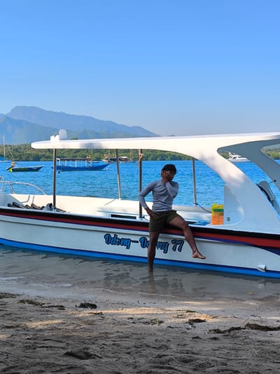 This image, captured in Gerokgak, Indonesia, displays a vibrant marine and coastal scene under a clear, light blue sky. In the foreground, a barefoot male individual with dark hair, wearing a light grey long-sleeved shirt and dark olive green shorts, poses leaning against the port side of a white speed boat. His right foot is submerged in the shallow water at the waterline, while his left lower leg rests on the boat's hull. His left hand is on his hip, and his right hand is raised to his cheek.
The boat, named "Odong - Dorong 77" in blue script on its hull, features blue and red/black stripes along its side. It is partially beached on a sandy shore, with its bow pointed towards the viewer's left. The boat is equipped with a white canopy supported by metal poles. Inside the boat, black fins are visible near the front. On the boat's deck, to the right of the man, a yellow plastic bag and an orange container are present.
The immediate foreground consists of wet and dry sand, showing footprints and scattered small patches of dark seaweed. Beyond the boat, clear blue water extends, dotted with several other vessels. These include a green and yellow long boat with a canopy to the far left, a longer blue boat with a canopy in the middle distance, and a large white yacht further out on the right. Additional boats are vaguely visible in the background.
The background features a distant landmass characterized by lush green forested hills and mountains, receding into a hazy blue. The sky above is entirely cloudless. The scene is illuminated by strong sunlight, casting defined shadows, suggesting a bright day.