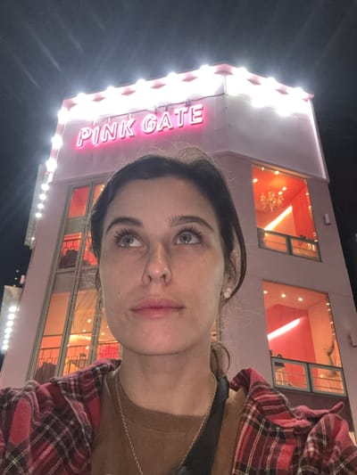 A low-angle, medium shot captures a young woman looking upwards and to the left with her face filling the foreground. She has fair skin, brown hair, and is wearing a red and black plaid shirt over a brown t-shirt and a silver necklace. Behind her, a brightly lit pink building with neon signage that reads "PINK GATE" dominates the background. The building has multiple floors with large windows showcasing pink interiors and what appears to be a chandelier in one of the upper windows. The image is taken at night, and the surrounding environment is dark, suggesting an urban setting. The location is identified as Busan, South Korea.