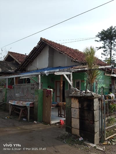 The image shows a small, green and white house with a wooden roof. The house is located in Anjatan, Indonesia, at coordinates -6.388 latitude and 107.93 longitude. There is a wooden fence in front of the house and a wooden table with a colorful tablecloth sitting in front of the fence. There is a palm tree growing next to the house. The ground is paved with concrete. The house appears to be in a state of disrepair, with some of the roof tiles missing. There are some items of clothing hanging on a clothesline in the background.