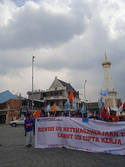 The image shows a protest in Yogyakarta, Indonesia, at the intersection of Jalan Jenderal Sudirman and Jalan Malioboro (-7.783, 110.367). A large group of people are holding a banner that reads "Revisi UU Ketenagakerjaan 13 Cabut UU Cipta Kerja" (Revision of Labor Law 13, Withdraw the Job Creation Law) in the foreground. The protesters are holding a variety of flags and banners, including a banner that says "BURUH BERSATU TIDAK BISA DIKALAHKAN" (Labor United Cannot Be Defeated). The protesters are standing in front of a building with a white facade, which is likely a government building. There is a tall, slender, white structure with a golden top behind the protesters. The sky is overcast with grey clouds. The scene is a street intersection. There is a car visible to the left of the image.