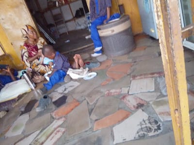 This image depicts an indoor scene in Hoche, Nigeria, likely within a commercial establishment or an open-fronted building. The floor is composed of irregularly shaped, mismatched concrete or tile pieces in shades of grey, orange, and white.

In the left foreground, a woman in a patterned dress and headwrap sits on the floor, holding an infant dressed in light blue. Beside her, a person lies reclined on a white mat, partially obscured, with a silver thermos flask nearby. In the center, a young boy in a blue plaid shirt and blue trousers sits on the floor, looking down at a white plate containing food. His bare feet rest on a white cloth, and a single dark shoe lies near him.

In the mid-ground, towards the right, another individual wearing blue trousers and a blue patterned shirt sits on a brown and beige ribbed plastic bin or cooler. Behind this person, a silver-grey refrigerator or freezer unit is visible against a yellowish-brown wall with peeling paint. Shelving with various items is present in the far left background. A prominent yellow wooden beam runs vertically in the right foreground, partially obscuring the view. The overall lighting suggests an indoor setting with ambient light.