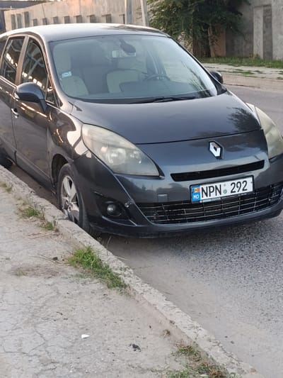 The image shows the front of a dark grey Renault Scenic car parked on the side of a street in Bălți, Moldova. The car is positioned at an angle, with its front wheels close to the curb. The license plate reads "NPN 292 MD". The surrounding environment includes a concrete curb, a dirt shoulder, and a paved road. In the background, there are buildings and trees. The lighting suggests it is daytime, possibly late afternoon, with warm light reflecting off the car and buildings. There are no people visible in the image. The car appears to be in normal condition, parked on a residential or commercial street.