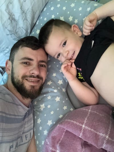 An adult male and a young child are depicted in a close-up selfie, positioned side-by-side, lying down on bedding. The scene is located in the city of Hadzhidimitrovo, Bulgaria. The adult male, positioned on the left foreground, has short dark hair, a dark full beard, and is smiling directly at the camera, revealing teeth. He wears a light-colored t-shirt with horizontal stripes, featuring the partially legible word "PACIFIC". The child, positioned to the right of the adult male and slightly further back, has short dark hair and is also smiling directly at the camera, revealing teeth. The child wears a black t-shirt with a partial light-colored graphic on the chest, and one hand is raised towards the mouth area. Bedding elements include a light grey sheet with a dark grey sailing ship pattern behind the adult male's head, a light blue pillow with white star patterns positioned between the two individuals, and a purple blanket with a white grid pattern of small squares and rectangles on the lower right. The lighting is soft and even across the faces of both subjects.