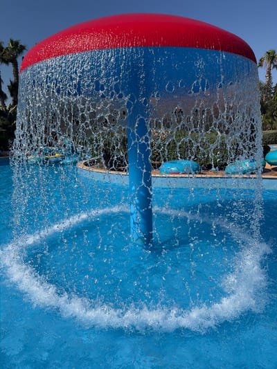 This is a close-up, rotated photograph taken at a water park in Trachoni, Cyprus. The primary subject is a large, spherical water feature, with a red band around its upper portion and a blue lower portion. Water cascades down from the top of this object, creating a textured curtain of droplets that obscure the background.
The background, visible through the falling water, shows a swimming pool with bright blue water. Further back, there are hints of palm trees and other greenery, suggesting an outdoor setting. The sky is a clear, bright blue, indicating a sunny day, likely midday given the lighting.
The water feature itself appears to be a decorative element within the water park, possibly a fountain or part of a slide. The way the water breaks and splashes creates dynamic patterns of light and shadow on the blue surface of the pool. There is a blue pipe-like structure extending from the water feature into the pool.
The image is oriented vertically, with the sky at the bottom and the water feature at the top of the frame. The focus is on the interaction between the water and the colored sphere, highlighting the movement and texture of the water. No people are visible in the frame. The overall impression is one of a bright, active, and refreshing water park environment.