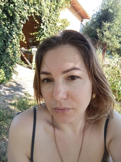 A woman with brown hair and brown eyes is captured in a close-up, outdoor selfie. She is wearing a dark-colored tank top and a gold necklace. The background consists of lush green foliage, a wooden structure resembling a shed or building, and the ground is covered with dry grass and dirt. The lighting suggests a sunny day. The exact location within Mykolaiv, Ukraine, cannot be determined from the provided image.
