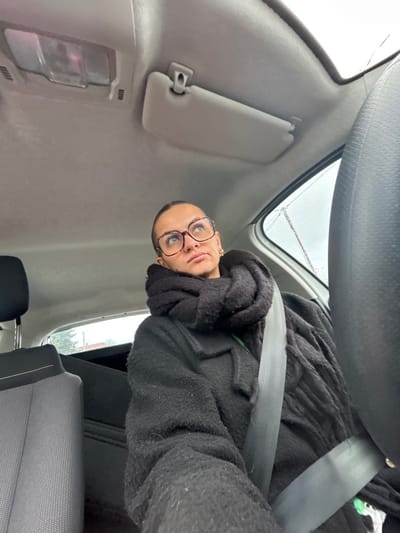 An individual is seated in the passenger front seat of a vehicle, described as being within the city of Saint-Cyprien, France. The subject, appearing to be female, wears large, rectangular eyeglasses with brown frames, a thick black scarf wrapped around her neck and shoulders, and a dark, textured long-sleeved garment, possibly a coat or sweater. Her hair is pulled back, and small gold hoop earrings are visible. She is secured by a grey seatbelt diagonal across her chest and lap, angled towards the left. Her head is tilted slightly back, and her eyes gaze upwards and to the left with a contemplative or pensive expression, indicated by slightly pursed lips.

The vehicle's interior features a light grey headliner. An overhead light fixture and a deployed grey sun visor are visible above the subject. To the left, a portion of an adjacent grey seat with black trim is visible, likely the driver's seat. Through the windows on the right side, an overcast sky with indistinct outdoor elements, possibly including power lines or rain streaks, is visible. The environment inside the vehicle is lit by ambient light, suggesting daylight conditions.
