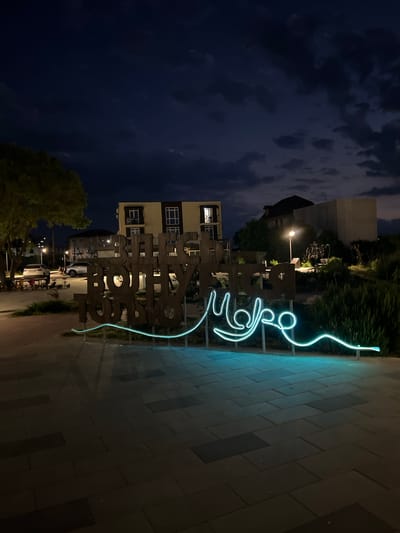 At nighttime in a public square in Shchyolkino, Ukraine, a large, illuminated neon sign spells out "BEING ON THE SEA" in Cyrillic script. The sign is composed of individual wooden letters, some of which are partially obscured by shadows. The neon light, a vibrant turquoise, outlines the words and extends in a wavy line below. The ground is paved with rectangular tiles, and a few streetlights cast a warm glow. In the background, a multi-story building with illuminated windows is visible, along with other structures and trees. The sky is dark, with some clouds visible.