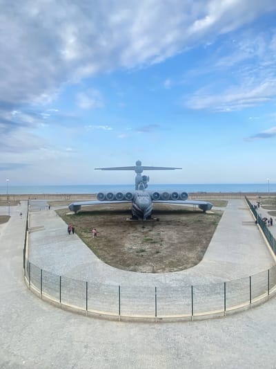 This image shows a large, gray amphibious aircraft, likely an Ekranoplan, on display outdoors. The aircraft is positioned centrally in the frame, with its wings extended and multiple engines visible along the wingspan. The environment appears to be a park or museum area near a body of water, suggested by the presence of a beach and the ocean in the background. The sky is partly cloudy with blue sky visible. Several people are scattered in the scene, walking on paved pathways and observing the aircraft. A green fence encloses parts of the area. The ground around the aircraft is covered with dry grass and dirt. The location is identified as Arablyar, Russia.