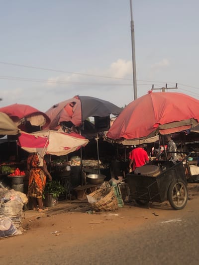 This image depicts a bustling outdoor street market scene, likely in Nsugbe, Nigeria, under a clear daytime sky with scattered clouds. The primary activity is market vending and shopping.

The scene is dominated by numerous large, colorful market umbrellas providing shade over makeshift stalls. These umbrellas, seen in shades of red, pink, white, and dark blue/grey, appear well-used, with some visible patches and wear. Beneath them, vendors are set up along an unpaved, dirt road. Various market stalls and carts are visible, including a prominent black wheeled cart.

Several people are present, though many are partially obscured by the market structures. In the lower right foreground, a woman wearing a vibrant orange and black patterned skirt is seen bending over, attending to her goods. Near her, baskets filled with red produce, possibly tomatoes or peppers, are arranged on the ground. Another person wearing a red shirt is partially visible behind a large red umbrella.

A utility pole with power lines extends into the background against the bright sky. The ground is an earthy, light brown color, consistent with an unpaved road or market area. The overall impression is one of a busy, functional market operating outdoors. No legible text is visible on any signs or objects within the frame.