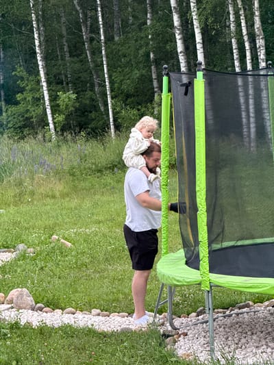 A Caucasian man, appearing to be in his late 20s or early 30s, is standing outdoors in Ryzhkovo, Russia, facing a trampoline. He has dark hair, a beard, and is wearing a white t-shirt and black shorts. A young Caucasian child, with blonde curly hair, is seated on the man's shoulders, facing away from the viewer and towards the trampoline. The child is wearing a cream-colored hooded sweatshirt. They are on a grassy area, with a gravel path lined with stones to their left. In the background, a dense forest of birch and other trees is visible. The trampoline has a green safety pad and a black net enclosure. The overall lighting suggests it is daytime, likely late afternoon.