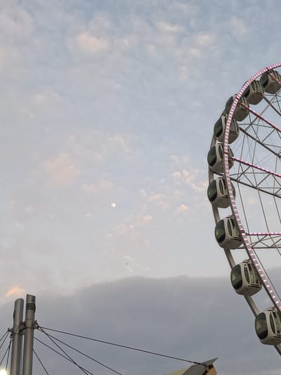 This image captures a view of a Ferris wheel in Guayaquil, Ecuador, with a hazy sky and clouds. The Ferris wheel, located in a city park, is partially visible on the right side of the frame, adorned with pink lights. The sky, a mix of light blue and grey, features scattered clouds and a faint glimpse of the moon. In the lower left quadrant, two metallic poles with suspended cables suggest infrastructure related to the park or an event. The overall atmosphere is subdued, possibly indicating dusk or an overcast day. No people are visible in the frame.