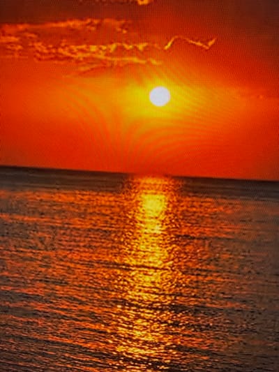 The image depicts a sunset over a body of water. The sky is a vibrant orange, with some hazy clouds visible. The sun, a bright white orb, is positioned in the upper center of the frame, casting a strong reflection on the water's surface. The water is textured with ripples, and the reflection of the sun creates a shimmering path of light extending from the horizon towards the viewer. The overall scene is one of natural beauty and tranquility. There is no discernible city or specific location identifiable within the image, and therefore, no connection to Szczecin, Poland can be made. The image contains no people, subjects, objects, actions, or interactions.