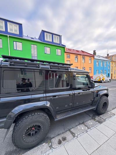 The image depicts a black off-road vehicle parked on a cobblestone street in Reykjavik, Iceland. The vehicle is oriented parallel to a row of brightly colored buildings in shades of blue, green, orange, and yellow. The sky above is overcast. A white commercial van is also parked further down the street. The reflection in the vehicle's windows shows a person holding a phone.
