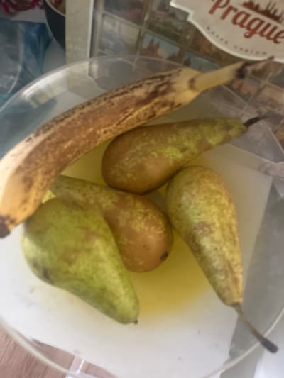 The image shows a close-up, slightly tilted view of a clear glass bowl containing fruit. The primary subjects are one ripe banana and four pears.
The banana, positioned on the left side of the bowl, is noticeably ripe with numerous dark brown spots on its yellow skin. Its stem is partially visible at the top. To the right of the banana, four pears are resting in the bowl. These pears are primarily greenish-yellow with some areas of reddish-brown mottling or spots, suggesting they are also ripe or ripening. Their stems are visible, pointing upwards or outwards from the fruit bodies. A pale yellow liquid or reflection appears to be at the bottom of the bowl, beneath the fruits.
In the blurred background, to the upper left, a white object with red text is partially visible. The word "Prague" is clearly discernible in a decorative red font, followed by "Water" and other faint, illegible text, suggesting it is a label or a sign, possibly for "Prague Water". The overall setting appears to be indoors, likely a kitchen or dining area, given the nature of the items. The lighting is bright and even, indicating daytime or a well-lit interior. The image provides no specific visual cues to confirm the location as Pokrovnik, Bulgaria.