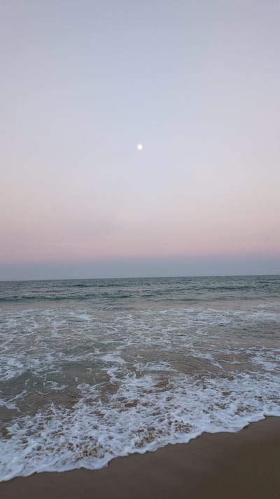 The image depicts a tranquil coastal scene, likely at dawn or dusk. The sky transitions from a pale gray to a soft pink, and a small, round object resembling the moon or a distant light source is visible. The ocean extends to the right, with waves breaking and creating foamy white patterns against the sandy beach. The scene appears devoid of people. The location is possibly a beach in Unknown, Vietnam.