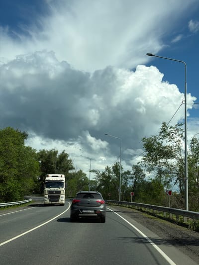 A gray SUV is driving on a curved asphalt road lined with a metal guardrail and green trees. The road is marked with a white line. The SUV has a license plate with Cyrillic characters. Ahead of the SUV, a white truck is also driving on the road. The truck has "BITRAK" written on its front grille. Traffic signs are visible along the roadside, including red arrow signs indicating a curve. The sky is filled with dramatic, large white and gray clouds, suggesting a dynamic weather system. Several streetlights are positioned along the road. The scene appears to be in a rural or semi-rural setting. The time of day is likely daytime, given the bright sky and sunlight casting shadows. The weather is overcast with patches of blue sky, indicating a mix of sun and clouds. The image was taken from a moving vehicle. No people are clearly visible. The location context provided is Верхнеозёрное, Russia.