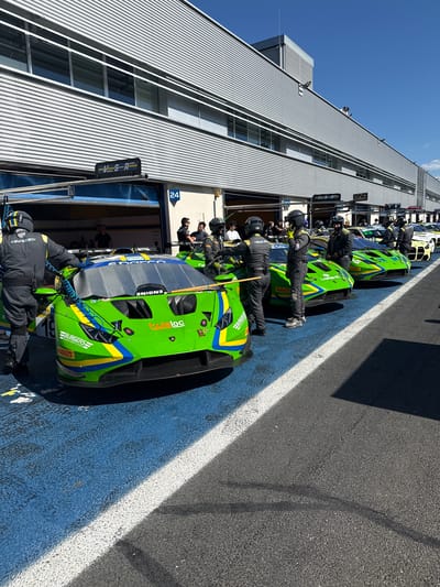 Here is a detailed description of the image:

**Scene Overview:**

The image shows a pit lane at what appears to be a racetrack or racing facility. Several race cars are lined up in their pit boxes, with teams of crew members attending to them. The time of day seems to be midday.

**Specific Elements:**

*   **Race Cars:** The prominent race car closest to the viewer is green with yellow and blue accents.
*   **Pit Crew:** Mechanics and team members wearing helmets are working on the cars.
*   **Pit Garage:** Each car is positioned in front of a pit garage, identified with a number "24".
*   **Buildings/Infrastructure:** A building with ribbed metal cladding is seen behind the pit garages.

**People:**

*   Crew members are present near the cars, likely performing maintenance, repairs, or adjustments.

**Actions/Interactions:**

*   Pit crews are working on their respective vehicles.

**Environment:**

*   Location: The image shows a pit lane, possibly a track. The V-S-R RACING sign is featured on the first pit garage.
*   Campagnano di Roma, Italy: This is the location.
